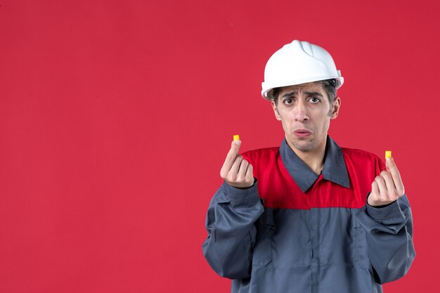 Vista frontal del joven trabajador molesto en uniforme con casco y sosteniendo tapones para los oídos en la pared roja aislada