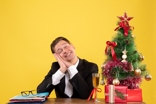 Vista frontal joven trabajador sentado con regalos de Navidad y árbol soñando