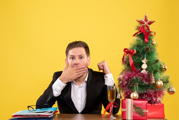 Vista frontal joven trabajador sentado con regalos de Navidad y árbol