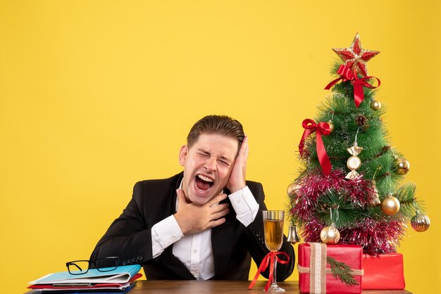 Vista frontal joven trabajador sentado con regalos de Navidad y árbol