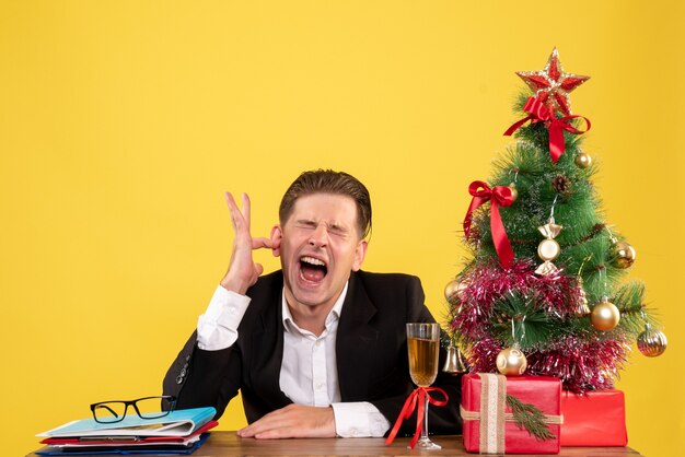 Vista frontal joven trabajador sentado con regalos de Navidad y árbol