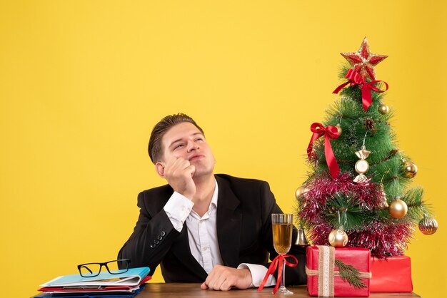 Vista frontal joven trabajador sentado con regalos de Navidad y árbol