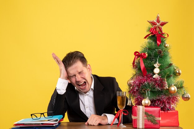 Vista frontal joven trabajador sentado con regalos de Navidad y árbol