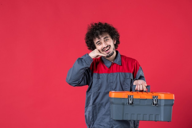 Vista frontal joven trabajador en uniforme con caja de herramientas sobre superficie roja