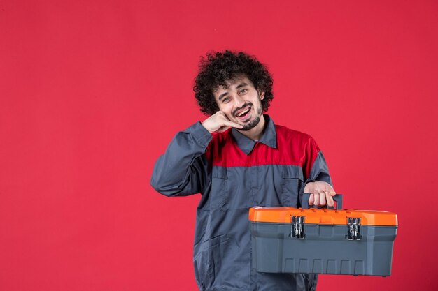 Vista frontal joven trabajador en uniforme con caja de herramientas sobre superficie roja