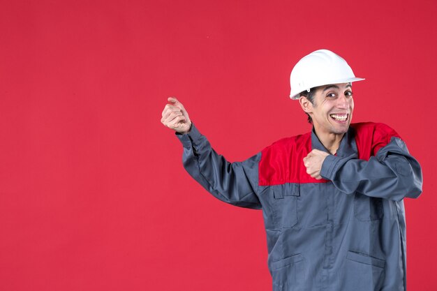 Vista frontal del joven trabajador en uniforme con casco y sentirse feliz en la pared roja aislada
