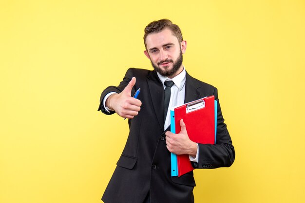 Vista frontal del joven en traje negro que parece satisfecho sosteniendo el portapapeles rojo y la carpeta azul haciendo el signo de ok mientras está de pie en amarillo