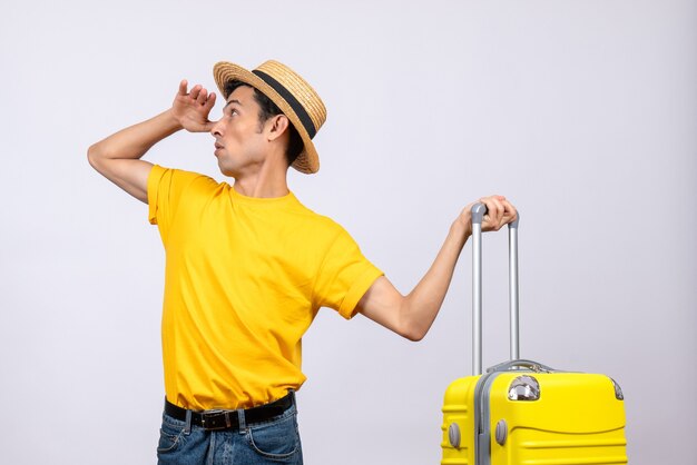 Vista frontal joven turista de pie cerca de la maleta amarilla mirando algo sosteniendo la maleta