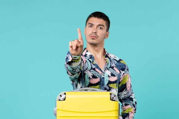 Vista frontal del joven varón preparándose para las vacaciones con bolsa amarilla mostrando su dedo en la pared azul