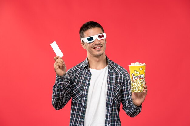 Vista frontal joven varón sosteniendo paquete de palomitas de maíz y boleto en la pared de color rojo claro cine cine cine película masculina