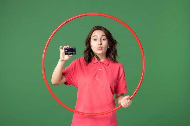 Vista frontal joven vendedora con hula hoop y tarjeta bancaria en superficie verde