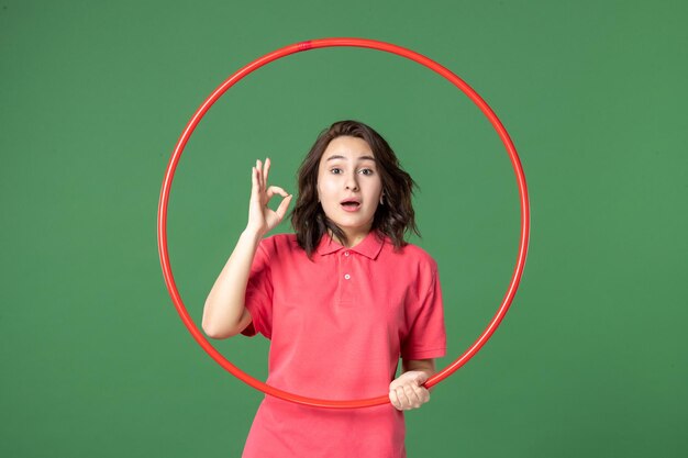 Vista frontal joven vendedora sosteniendo hula hoop rojo sobre fondo verde trabajo uniforme de venta trabajo de atleta de compras