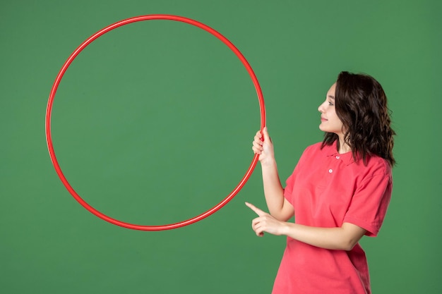 Vista frontal joven vendedora sosteniendo hula hoop rojo sobre superficie verde