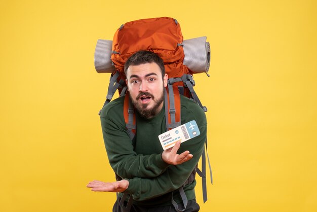 Vista frontal del joven viajero confundido con mochila y mostrando el boleto sobre fondo amarillo