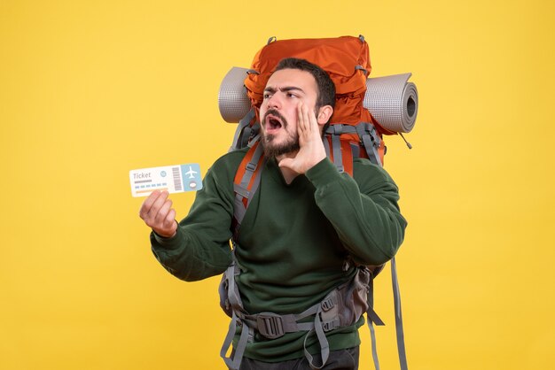 Vista frontal del joven viajero enojado con mochila y sosteniendo el boleto llamando a alguien sobre fondo amarillo