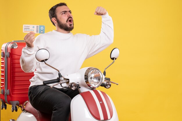 Vista frontal del joven viajero orgulloso sentado en la motocicleta con la maleta sosteniendo el boleto sobre fondo amarillo aislado