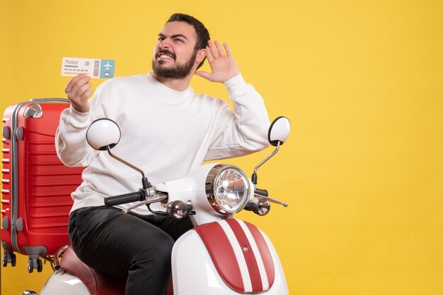 Vista frontal del joven viajero sonriente sentado en una motocicleta con la maleta sujetando el boleto escuchando los últimos chismes sobre fondo amarillo aislado