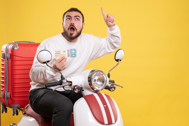 Vista frontal del joven viajero sorprendido sentado en la motocicleta con la maleta sosteniendo el boleto apuntando hacia arriba sobre fondo amarillo aislado