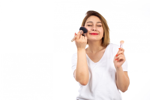 Una vista frontal jovencita con camisa blanca y pantalones modernos ligeros haciendo maquillaje en el blanco