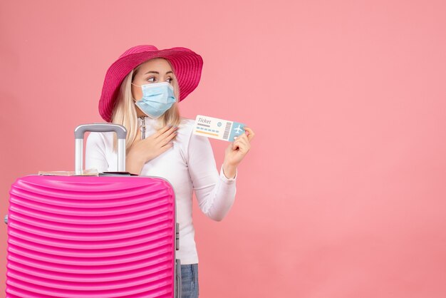 Vista frontal jovencita sosteniendo billete de avión de pie cerca de la maleta