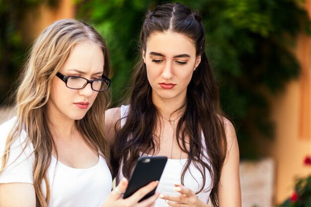 Vista frontal de jóvenes hermosas chicas mirando algo en el teléfono con caras serias al aire libre en el día caluroso