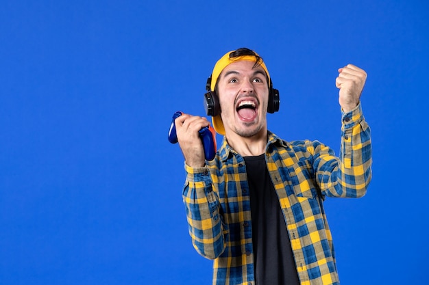 Vista frontal del jugador masculino emocional con gamepad jugando videojuegos en la pared azul