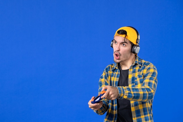 Foto gratuita vista frontal del jugador masculino con gamepad y auriculares jugando videojuegos en la pared azul
