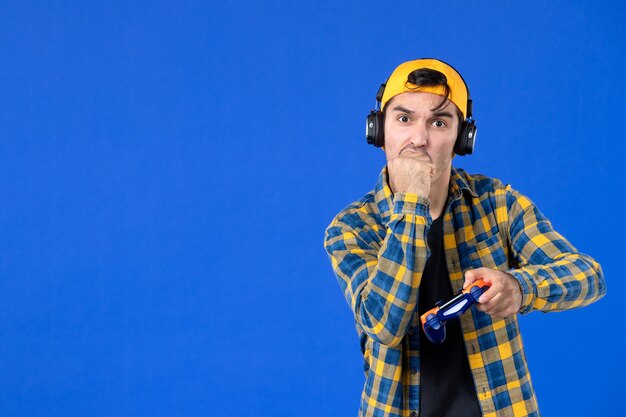 Vista frontal del jugador masculino con gamepad y auriculares jugando videojuegos en la pared azul