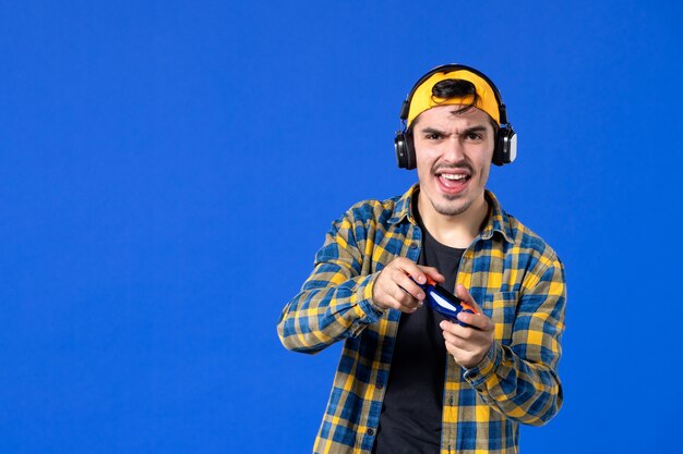 Vista frontal del jugador masculino con gamepad y auriculares jugando videojuegos en la pared azul