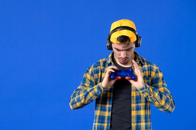 Foto gratuita vista frontal del jugador masculino con gamepad y auriculares jugando videojuegos en la pared azul