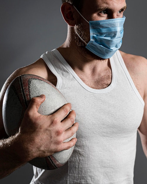 Vista frontal del jugador de rugby masculino atlético con máscara médica y sosteniendo la pelota