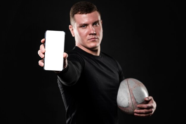 Vista frontal del jugador de rugby masculino sosteniendo la bola y el teléfono inteligente