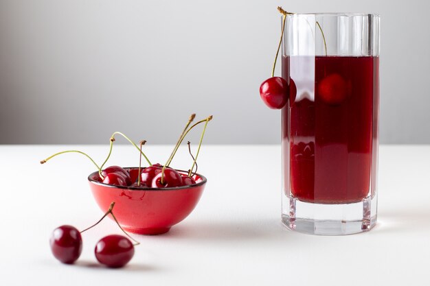 Vista frontal del jugo de cereza dentro de un vaso largo con cerezas frescas en la superficie blanca