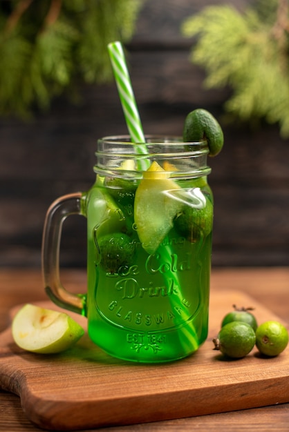 Vista frontal de jugo de fruta fresca en un vaso servido con tubos y feijoas de limones de manzana sobre una tabla de cortar de madera