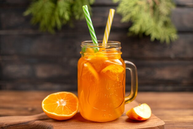 Vista frontal de jugo de fruta fresca en un vaso servido con tubos y manzana y naranja sobre una tabla de cortar de madera sobre una mesa marrón