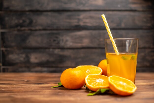 Vista frontal de jugo de naranja fresco en un vaso servido con un tubo de menta y naranjas enteras cortadas en el lado izquierdo sobre una mesa de madera