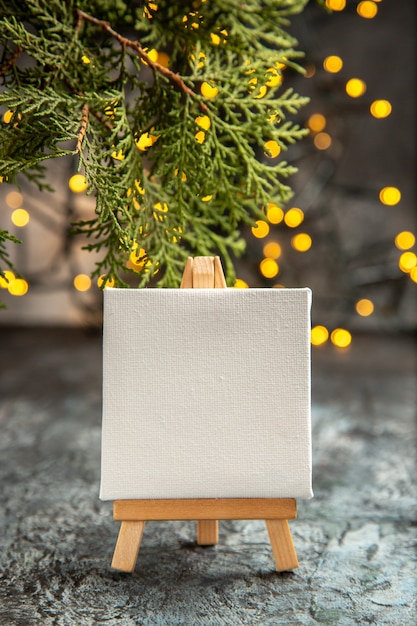Vista frontal lienzo blanco sobre caballete de madera luces de Navidad ramas de pino en la oscuridad