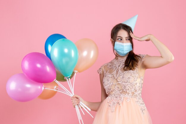 Vista frontal linda chica fiestera con gorra de fiesta sosteniendo globos de colores poniendo la mano en la sien