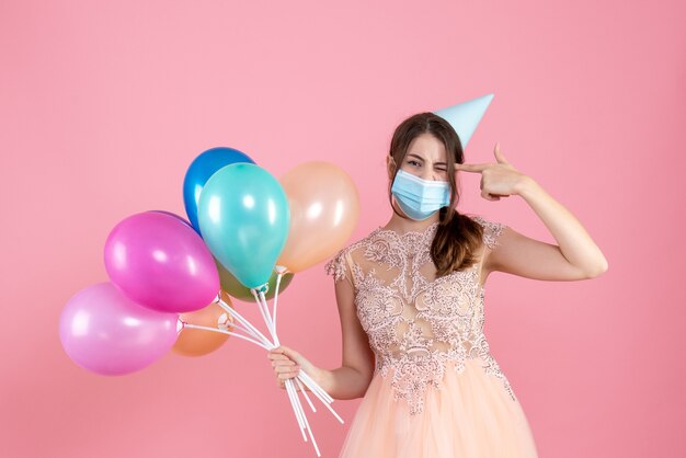 Vista frontal linda chica fiestera con gorra de fiesta sosteniendo globos de colores poniendo pistola de dedo en su sien