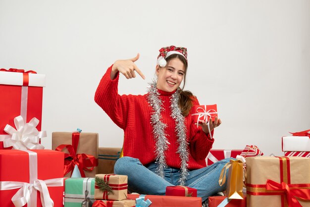 Vista frontal linda chica fiestera con gorro de Papá Noel apuntando a su presente sentado alrededor de regalos