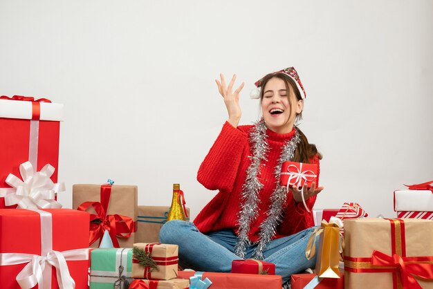 Vista frontal linda chica fiestera con gorro de Papá Noel sosteniendo presente y riendo sentado alrededor de regalos