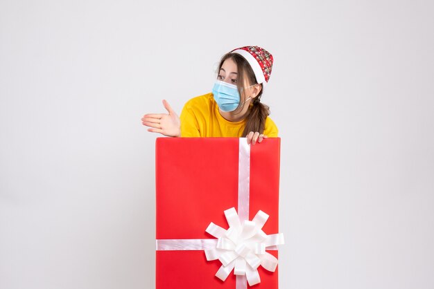 Vista frontal linda chica con gorro de Papá Noel dando mano de pie detrás de gran regalo de Navidad