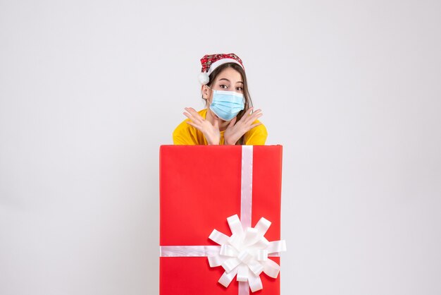 Vista frontal linda chica con gorro de Papá Noel y máscara de pie detrás de un gran regalo de Navidad