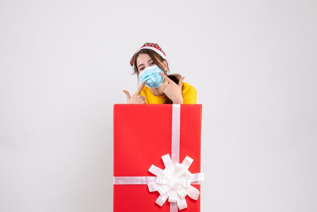 Vista frontal linda chica de Navidad con gorro de Papá Noel apuntando a su máscara de pie detrás de un gran regalo de Navidad