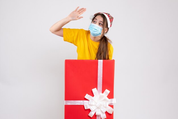 Vista frontal linda chica de Navidad con gorro de Papá Noel saludando a alguien de pie detrás de un gran regalo de Navidad