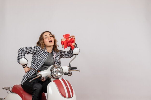 Vista frontal de la linda joven en ciclomotor con regalo y tarjeta en pared gris