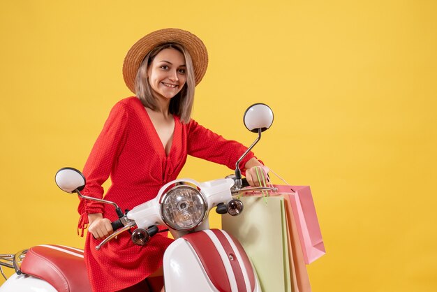 Vista frontal de la linda mujer joven en vestido rojo en ciclomotor sosteniendo bolsas de la compra.