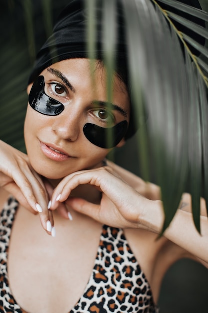 Vista frontal de una linda mujer con parches en los ojos debajo de la palmera. Hermosa modelo femenina posando sobre fondo tropical.