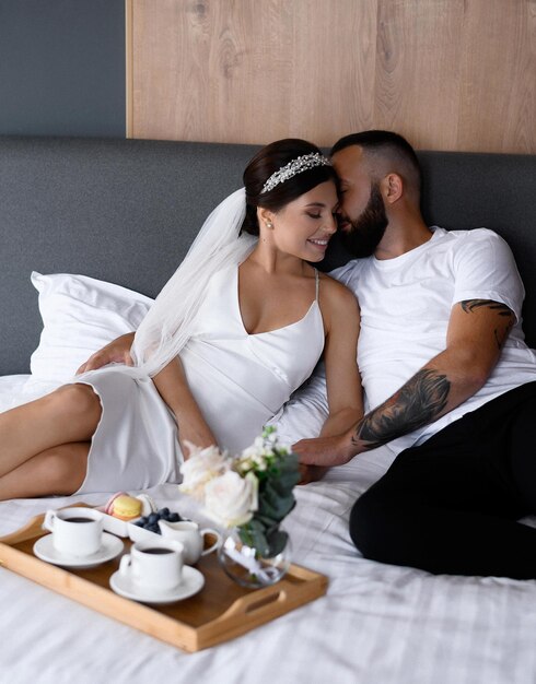 Vista frontal de la linda novia y el novio acostados juntos en la cama y tocándose durante la mañana de la boda Encantadora pareja relajándose en la cama durante el desayuno de la boda