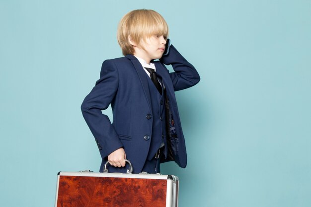 Una vista frontal lindo chico de negocios en traje clásico azul posando sosteniendo una maleta marrón-plateada hablando por teléfono moda de trabajo comercial